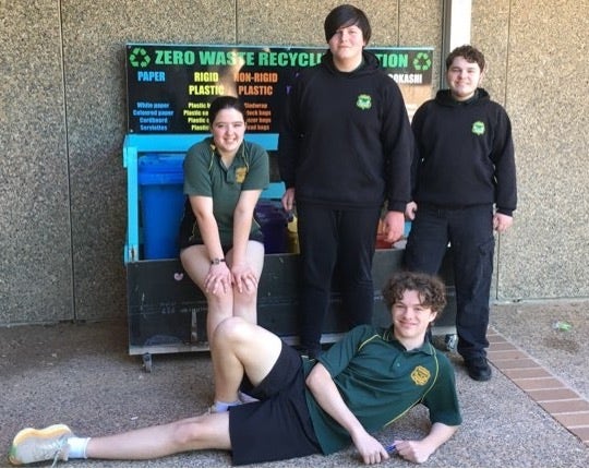 Four students stand in front of a wooden compartment that holds different types of bin for different categories of waste.