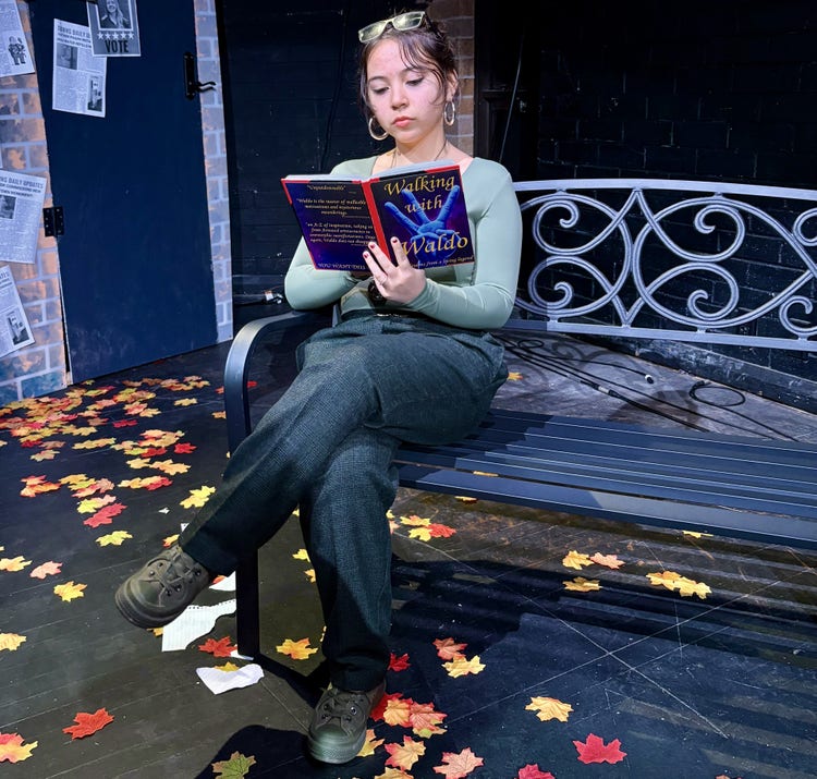 A female student is in a performance. She sits on a bench holding a book. She has a green shirt on and black pants and a pair of sunglasses on her head.