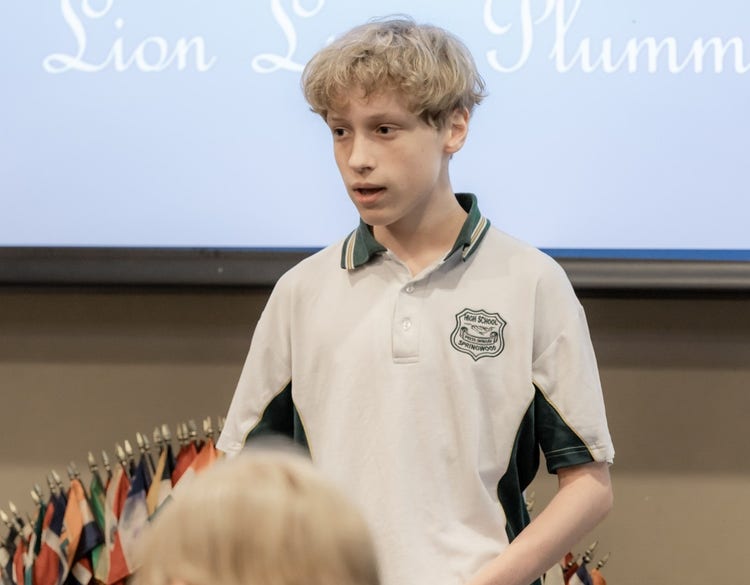 A blond haired male student stands in front of a crowd presenting his argument.